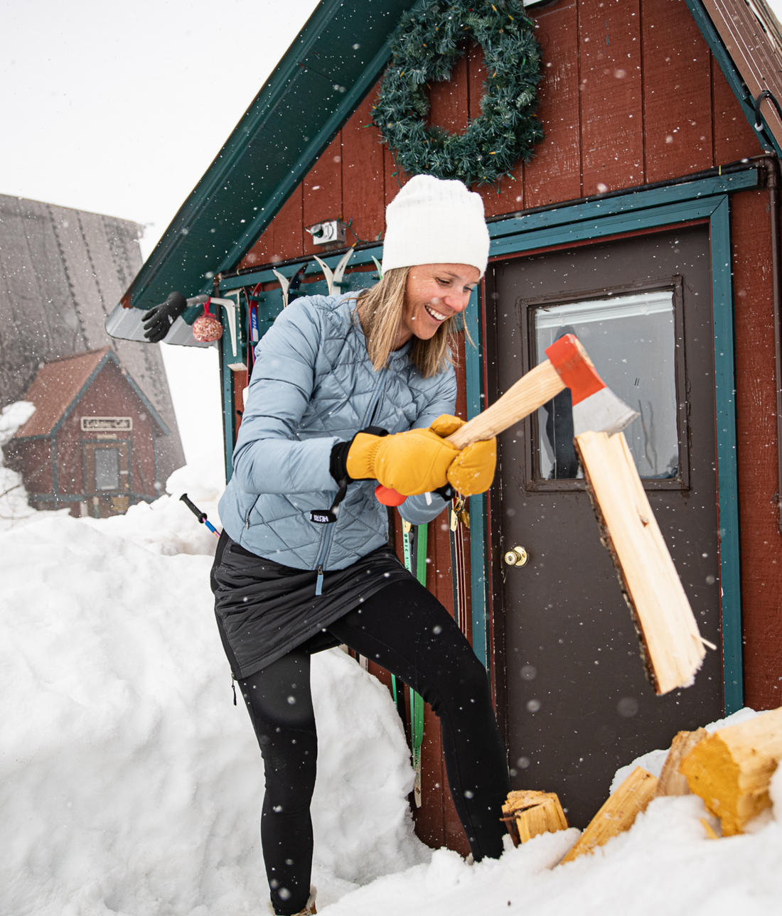 woman chopping wood outside snowy cabin in skhoop outfit
